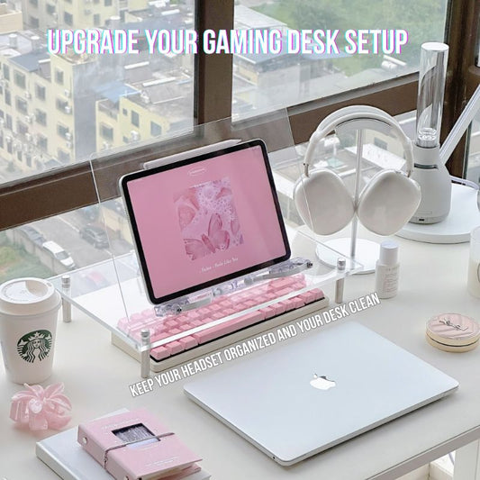 Aesthetic pink and white gaming desk setup with an iPad, laptop, pink keyboard, accessories, and wireless headphones displayed on the Aluminum Gaming Headphone Stand by a large window overlooking city views.