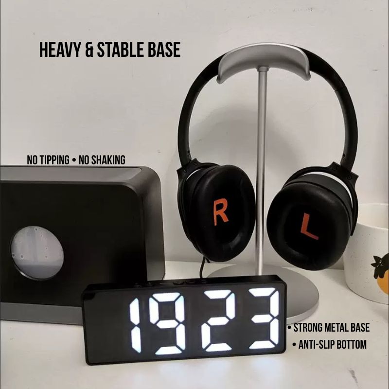 The Aluminum Gaming Headphone Stand with a heavy, anti-slip metal base holds black R/L-marked headphones. Nearby are a digital clock at 19:23, a black box, and a mug—perfect for any PC or gaming setup.