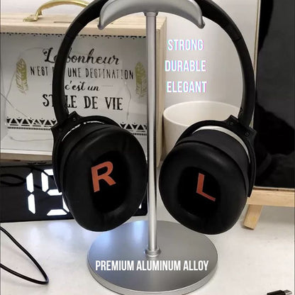 A pair of black headphones with bold red L and R labels rests on the Aluminum Gaming Headphone Stand, Heavy Base Desk Headset Holder for PC & Gaming Setup. "STRONG DURABLE ELEGANT PREMIUM ALUMINUM ALLOY" is shown. A digital clock and decor are behind.